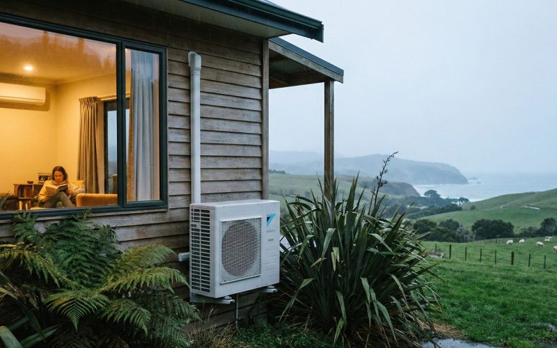 Cozy wooden hut with person reading indoors, overlooking green hills and ocean, with outdoor air conditioner and lush plants in foreground.